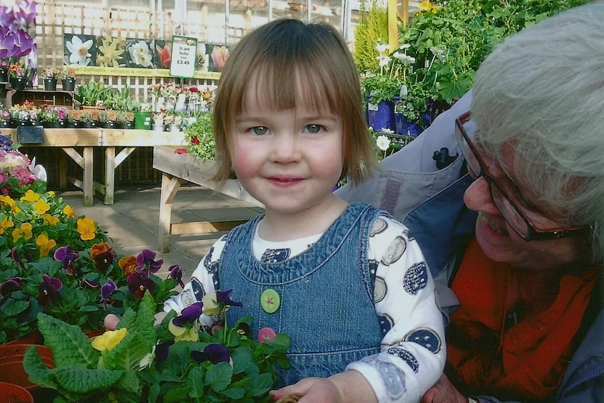 A toddler standing next to some plants at a garden center accompanied by her Granny