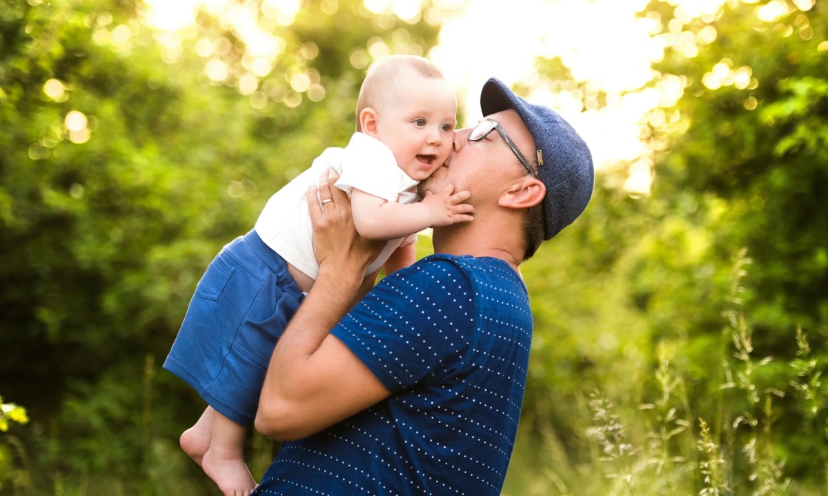 A father lifting his baby up, they are outside in front of some bright green trees